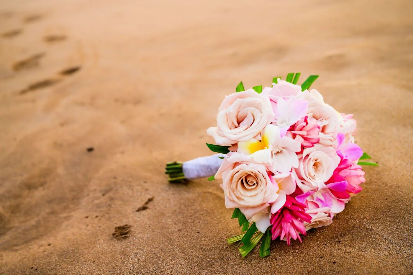 A blush and pink rose bridal bouquet resting on golden beach sand.