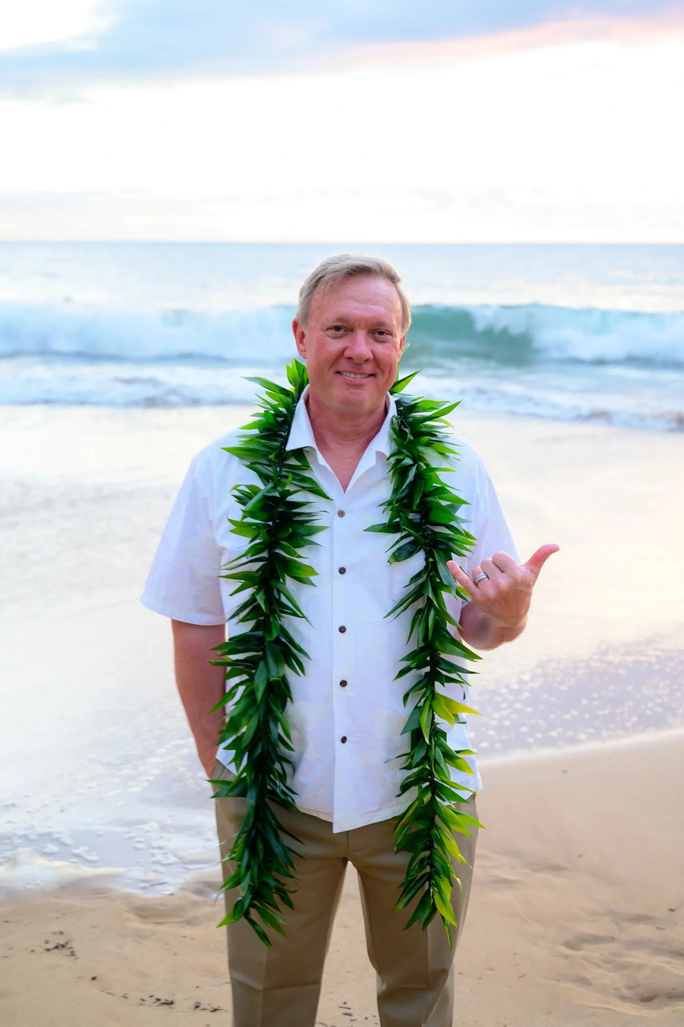 A man in a white shirt on the beach wearing a green ti-leaf maile lei.