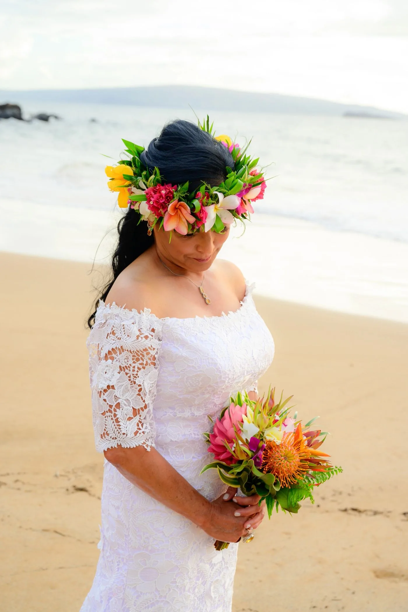 A bride in a white lace dress on the beach, wearing a tropical haku flower crown and holding a bouquet of protea, pincushion and orchids.