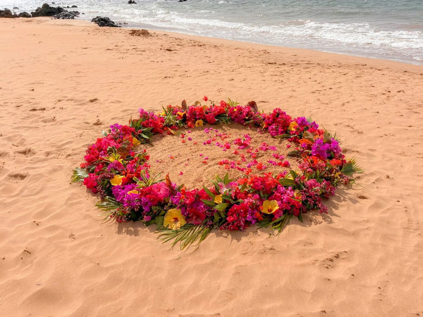 A vivid ring of hibiscus and bougainvillea flowers arranged on the sand near the shoreline.