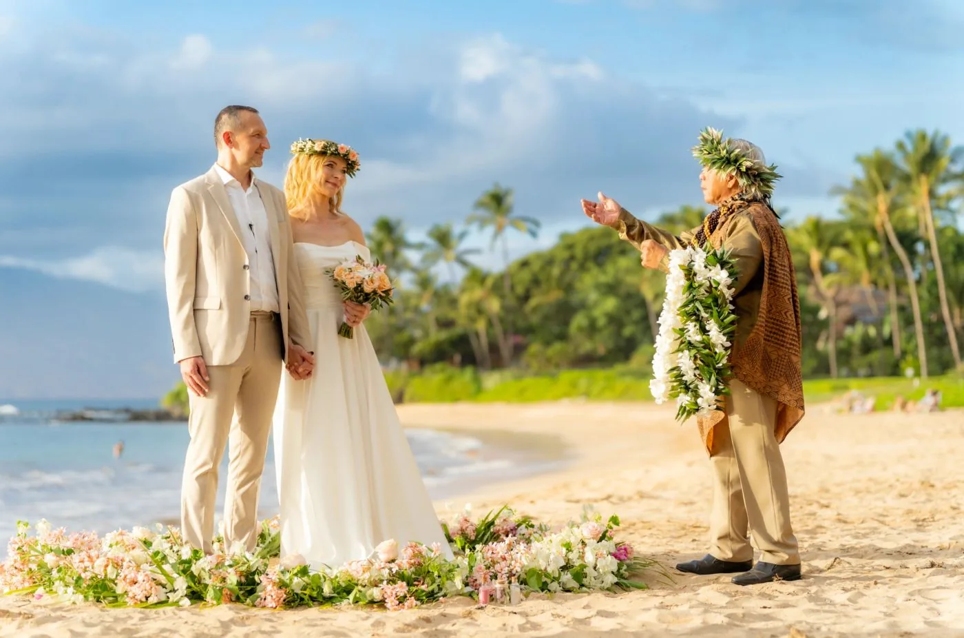 A bride and groom with a Hawaiian officiant during a beach ceremony inside a circle of pink and white flowers.