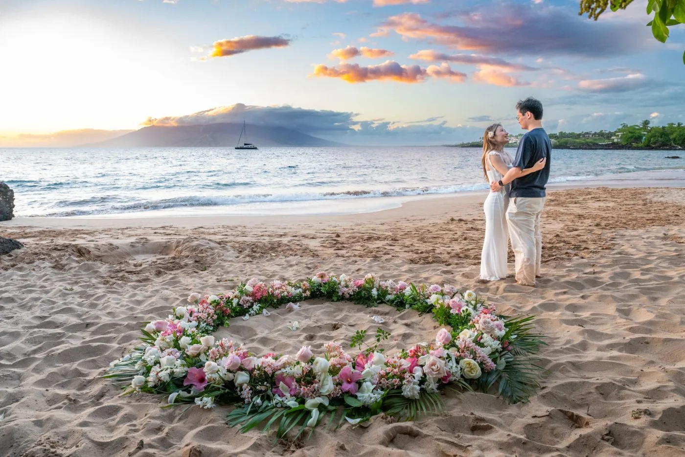 A couple embraces at sunset on a South Maui beach beside a circle of pink and white flowers arranged on the sand, with the West Maui mountains and a sailboat in the distance.