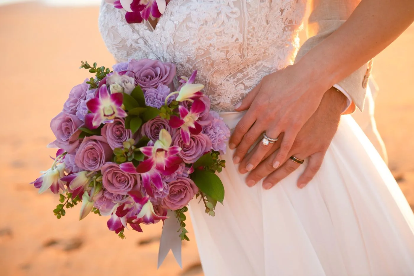 A lavender rose and magenta orchid bridal bouquet held by a bride, hands with wedding rings resting on her gown.